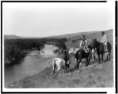 [Three Piegan Indians and four horses on hill above river], 1910